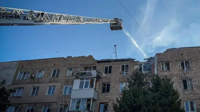 Firefighters on a crane spray water onto the top of a damaged building