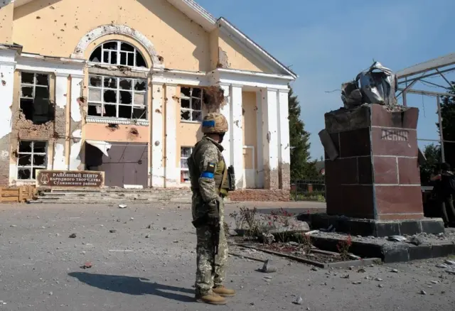 A Ukrainian serviceman stands near a damaged building in the city center of Sudzha,