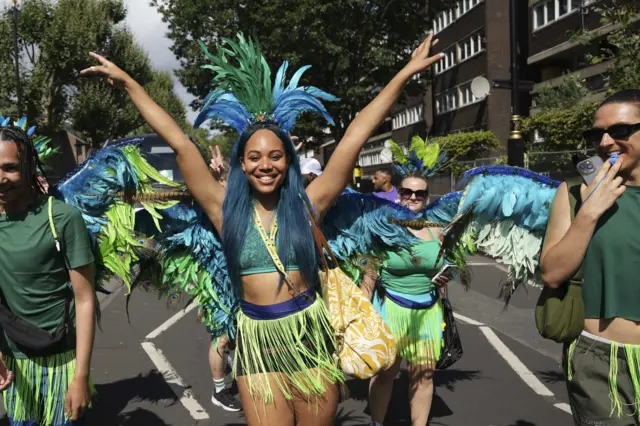 Notting Hill Carnival attendee wearing green and blue outit - shorts with green fringe and blue and green feather on her head and arms above her head as others stand nearby