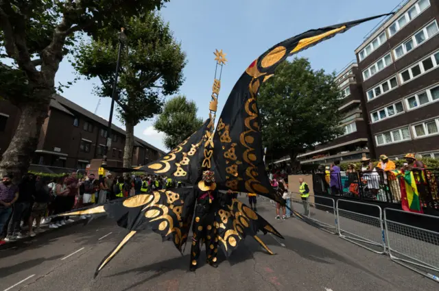 A person in a huge black and gold costume walking down an empty road at Notting Hill Carnival, there are people on each pavement watching.