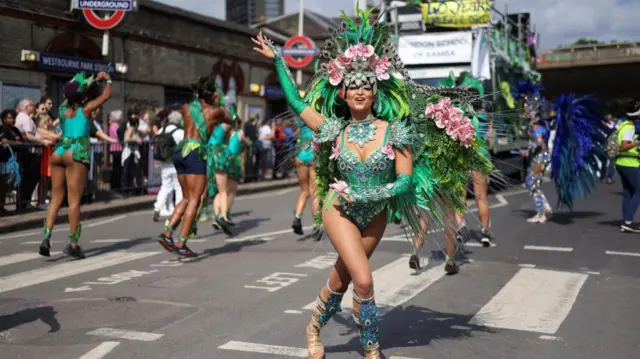 Woman in green Carnival outfit with feathers and pink flower headpiece outside Westbourne Park Tube station with parade van driving beind