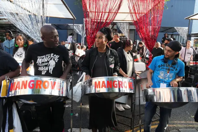 Three people stood next to each other with steel drums, two of which on the left and middle say Mangrove, as part of the Members of Mangrove Steelband who performed during Notting Hill Carnival's Panorama evening