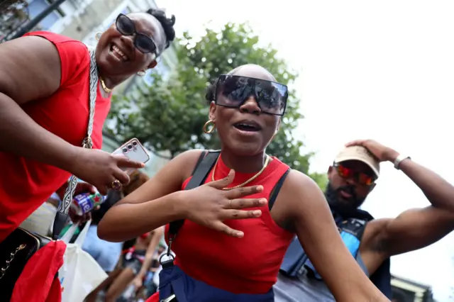 A woman dancing in a read top and sunglasses. There is a woman next to her, also in a red top ans sunglasses, and behind is a man with his hand on his head