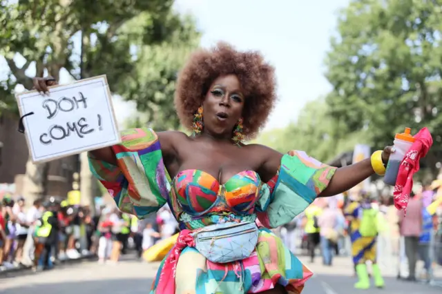 A woman dancing in the street in a brightly-coloured outfit. She is holding a sign saying "Doh come!"