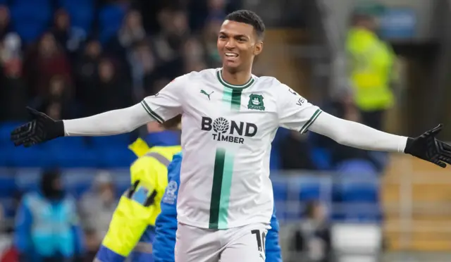 Morgan Whittaker, wearing Plymouth's white away kit with dark and lime green vertical strips down the front, smiles and stretches both arms out to his side as he celebrates a goal