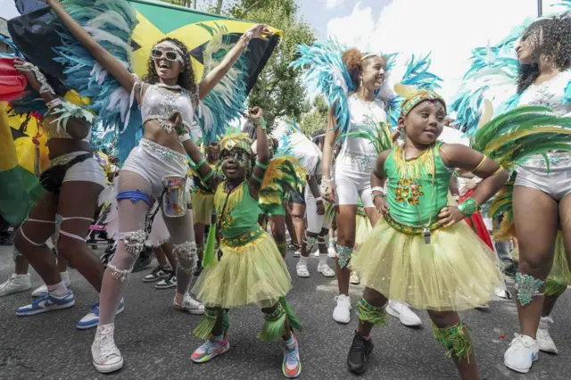 Two young girls in green tops, yellow tutus and wings made from black green and yellow feathers dancing alongside older girls dressed in white at Notting Hill Carnival, as one girl holds a Jamaican flag above her head