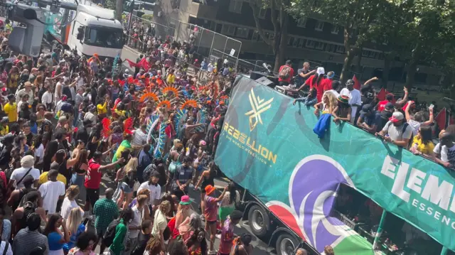 Crowds gathered in street at Notting Hill Carnival in bright coloured outfits as bus drives past full will revellers