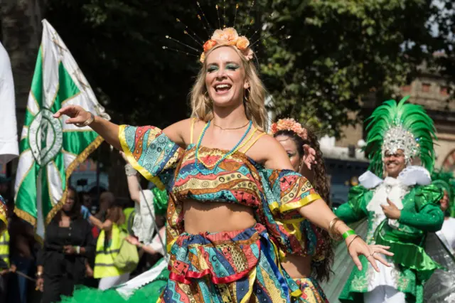 A samba performer in a brightly-coloured dress dancing, She has men dressed in green behind her