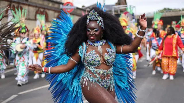 Woman in blue feathered dance outfit at Carnival with jewelled headpiece as more people in carnival outfits (blurred) walk behind her