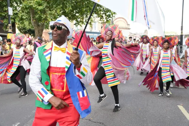 Man wearing multi coloured suit with yellow bowties holding umbrella in front of a group of girls wearing multicoloured striped dresses, capes and elaborate headpieces in parade through Notting Hill