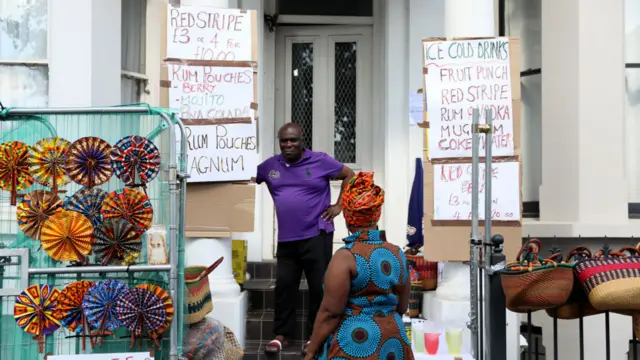 Man stood outside home in Notting Hill talking to woman facing away from camera with signs selling cold drinks and alcohol as well as rail of handbags and fans