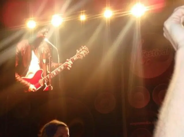 Noel Gallagher on stage at V Festival with leather brown jacket and red electric guitar with lights from the stage and arm of audience member in view