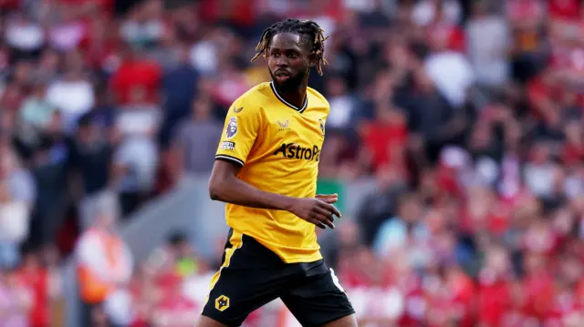 Boubacar Traore looks to his right and has his right arm across him as he half turns to look at something out of the picture. He is wearing Wolves' old gold home kit and black shorts. Behind him is a blurred background of fans wearing red.