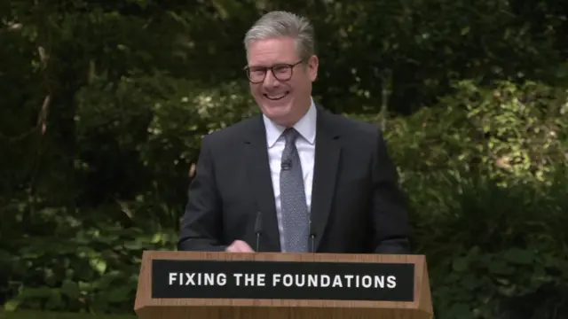 Keir Starmer laughing at podium while giving speech with 'fixing the foundations' slogan on his podium