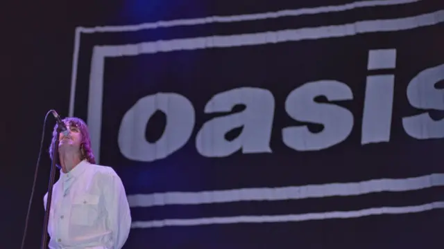 Liam Gallagher in white suit performing at Knebworth with microphone on stand and large banner reading Oasis with black background and white letters behind him