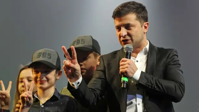 Zelenksy on stage with three children, all of them are holding up their middle and index fingers in a "v for victory" sign. Two boys are wearing hats which have #95 written on them