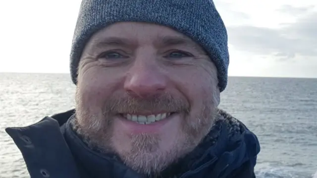 A selfie of Howard Irwin. He is by the sea and wearing a hat