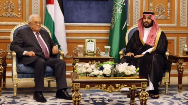 Mahmoud Abbas sitting in a chair next to Mohammed bin Salman, the Palestinian and Saudi Arabian flags are placed behind them