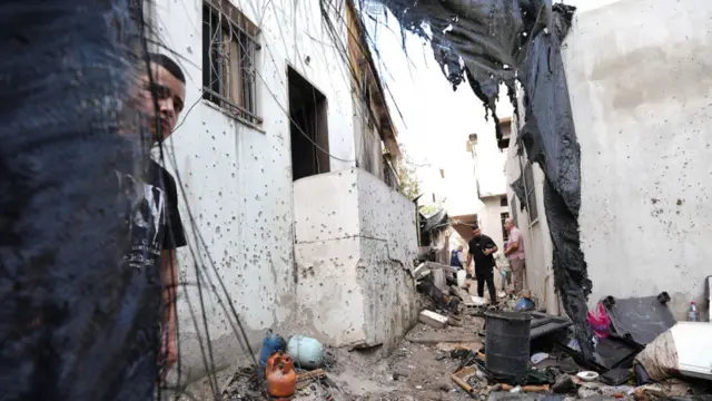 People stood in street covered in debris, walls on either side of narrow path covered in holes, as boy stands partly covered by damaged fabric as two men stand further back looking at the damage