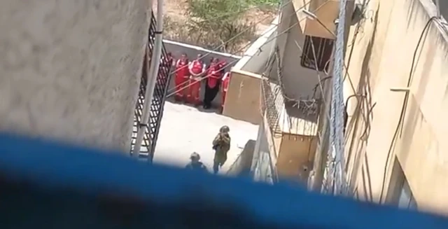 Five ambulance worker stand next to a wall, with two Israeli soldiers in uniform standing near to them