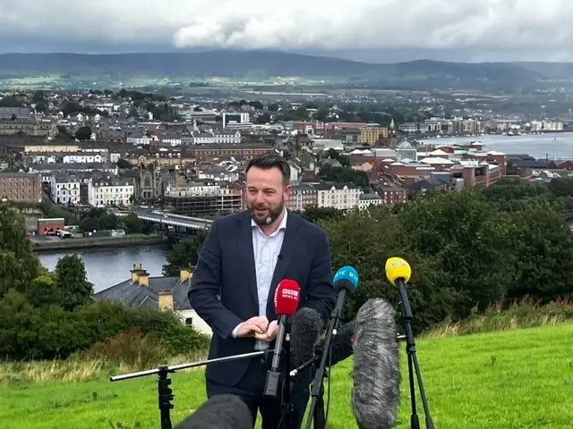 Colum Eastwood, wearing a suit, stands in front of microphones