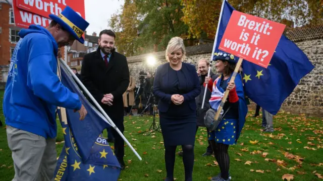 Colum Eastwood and Michelle O'Neill on college green in London with anti-Brexit protestors holding up signs