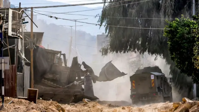 An excavator digs up the road, a second military vehicle in the background