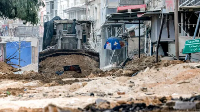 An excavator digging up a road, buildings and rubble to the right