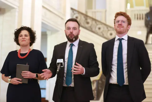 Colum Eastwood standing in the assembly flanked by Claire Hanna and Matthew O'Toole