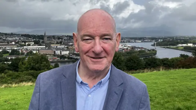 A man stands on a hill, wearing a grey suit with a light blue shirt