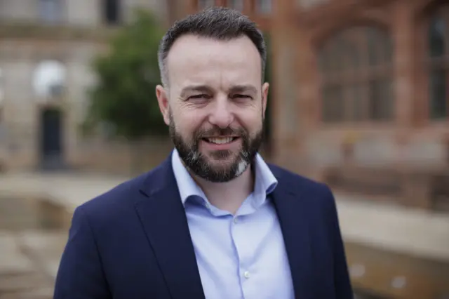 A man with a black beard and black hair, he is wearing a blue shirt and navy blazer