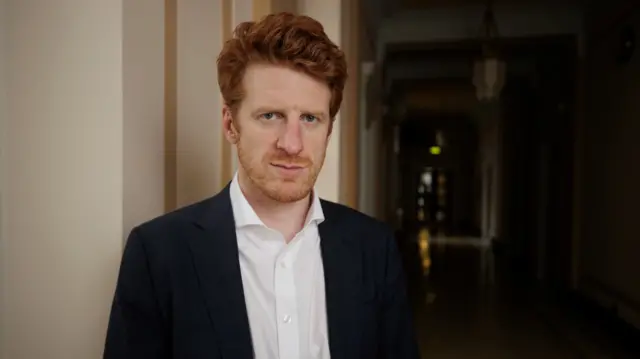 Matthew O'Toole standing in a corridor looking at the camera with a slight smile. He is wearing a suit with no tie