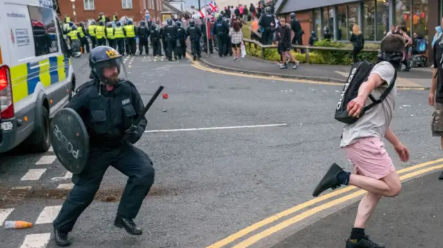 police with shield and baton chases a man who is wearing pink shorts