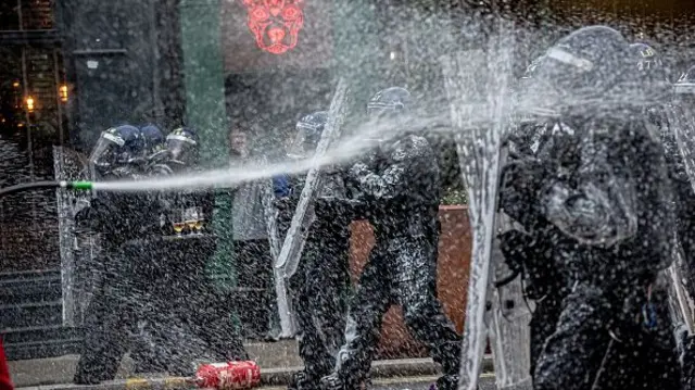 Officers in riot gear being sprayed by a water hose.