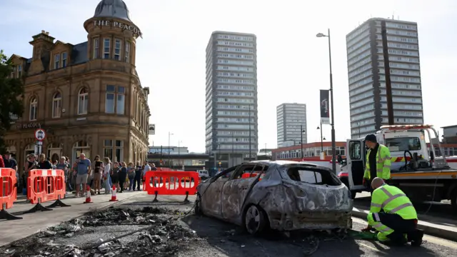 A burnt car is removed after a night of violent anti-immigrant demonstrations