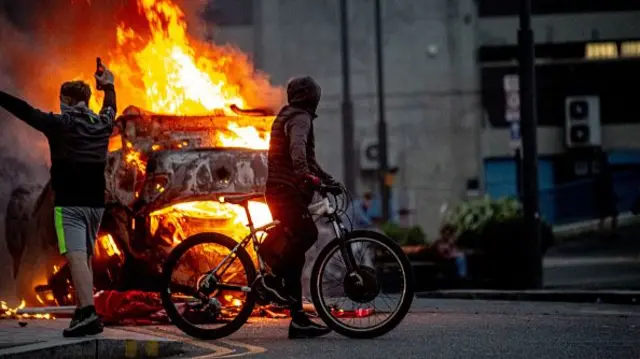 A man on bike in front of a vehicle on fire.