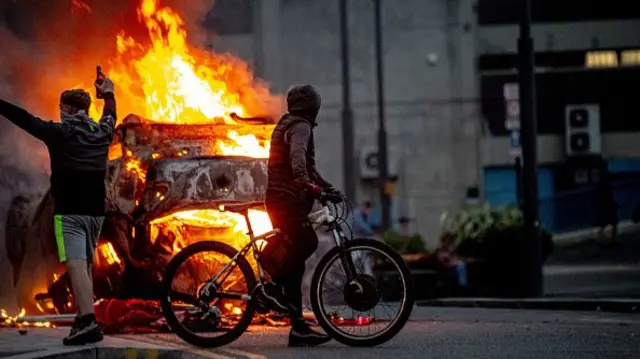 A man on a bike in front of a vehicle on fire.