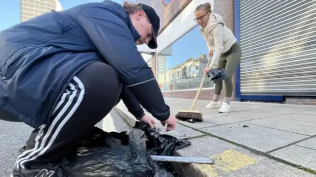 A man cleaning up a pavement, with a woman sweeping rubbish in background.