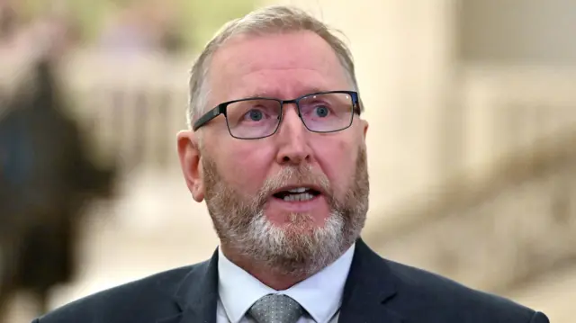 White haired man with a beard wearing navy glasses and dressed in a dark blue suit