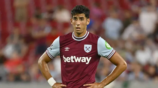 Nayef Aguerd, wearing West Ham's claret and blue home kit, puts his hands on his hips and looks at something off camera. Behind him are the blurred images of fans in the stand