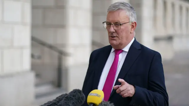 White haired man with grey glasses wearing a dark blue suit and hot pink tie standing in front of microphones