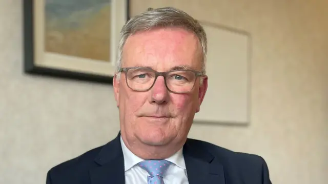 White haired man wearing grey glasses and dressed in a dark blue suit in a cream room