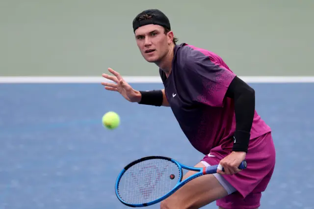 Jack Draper plays a backhand volley wearing a dark faded black and pink top with red-pink shorts, black undersleeve on his left arm, black baseball cap worn backwards and a black sweatband on his right wrist