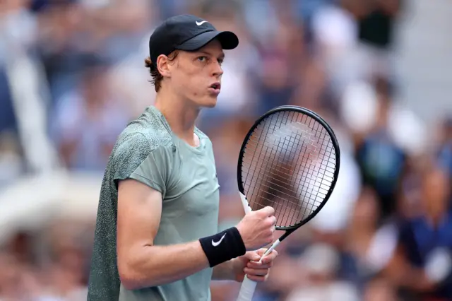 Jannik Sinner clenches his fist in celebration wearing a a grey Nike top, black baseball cap and black sweatband on his right wrist