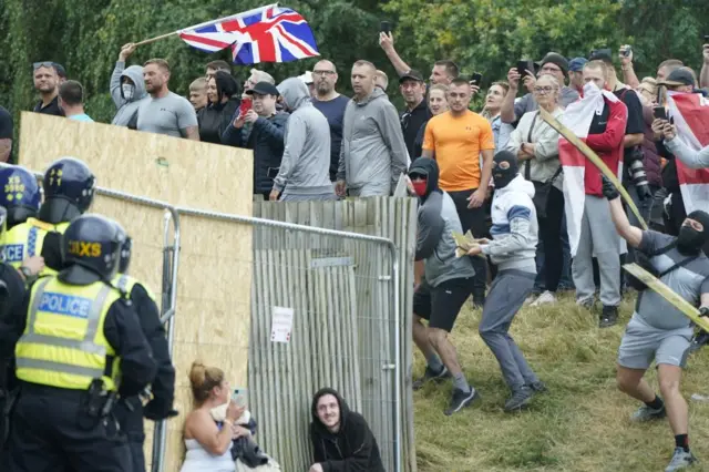 A large group of anti-immigration protesters facing police, two people are crouched, hiding.