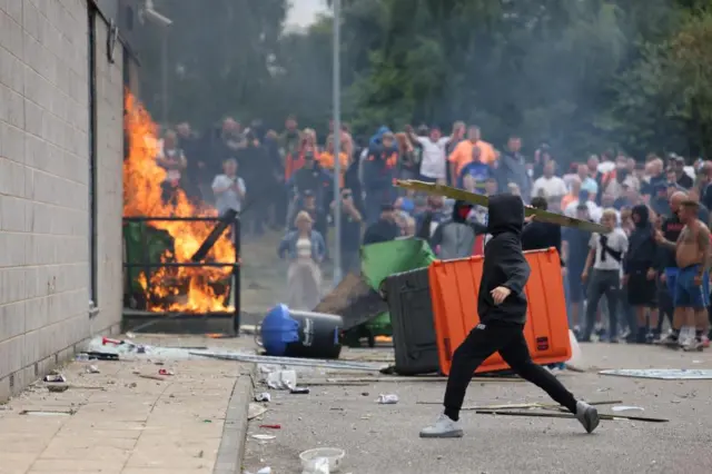 A small fire at a hotel, a man stands holding a fence pole like a javelin