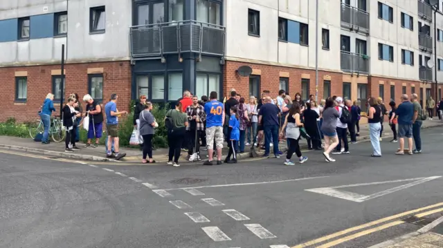 A large group of people stood on a street corner, in front of a block of flats