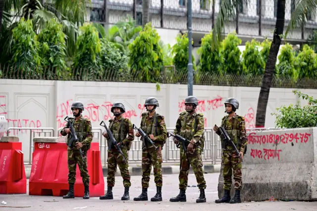 Armed soldiers carrying rifles stand guard at a roadblock, in front of a graffitied wall.