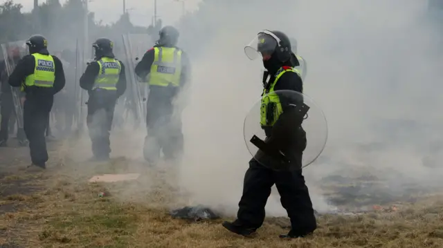 A police officer in riot gear walks with smoke and unrest in the background during a violent demonstration