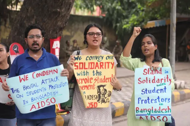 THree students holding protest signs in front of a row of trees.